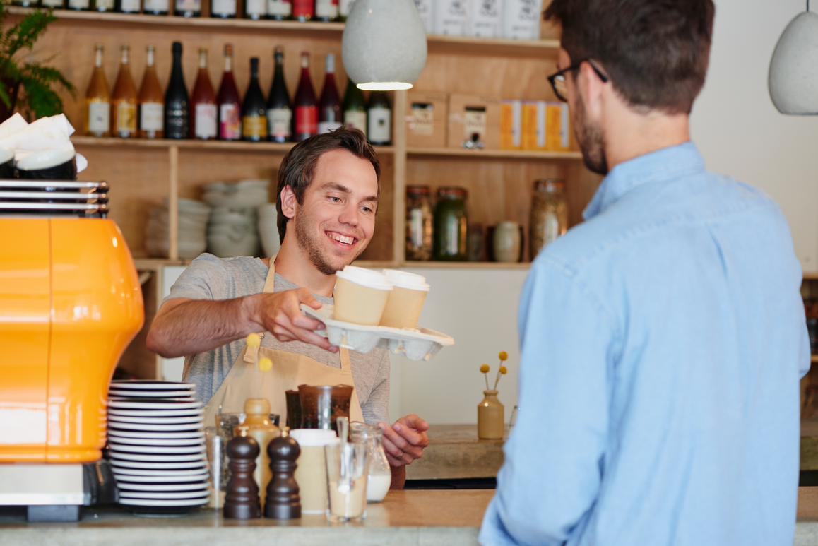 Een glimlachende cafémedewerker geeft een dienblad met twee kartonnen koffiebekers aan een formeel geklede klant.